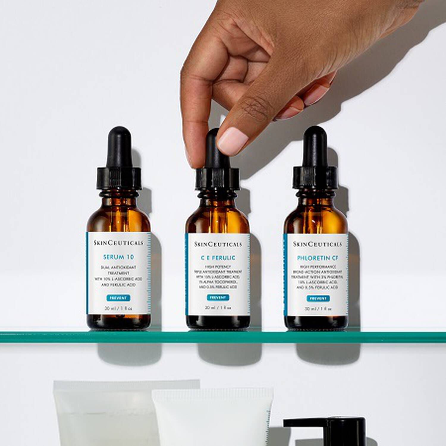 Main dans un flacon de serum, trois bouteilles de soins de la peau SkinCeuticals sur une etagere blanche.
