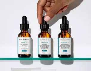 Main dans un flacon de serum, trois bouteilles de soins de la peau SkinCeuticals sur une etagere blanche.