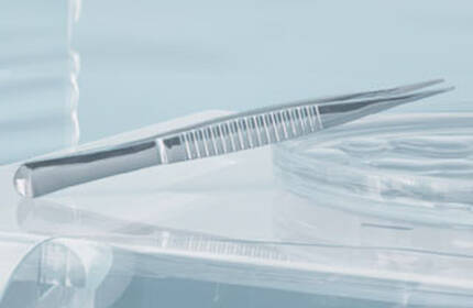 A close-up view of a dental or medical instrument, specifically a stainless steel tweezers, resting on a clear surface, with soft lighting and blurred background.