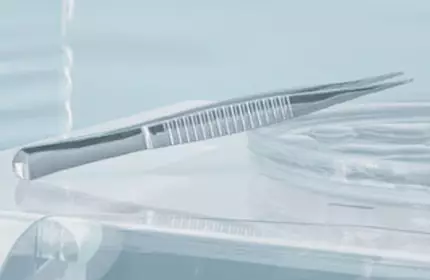 A close-up view of a dental or medical instrument, specifically a stainless steel tweezers, resting on a clear surface, with soft lighting and blurred background.