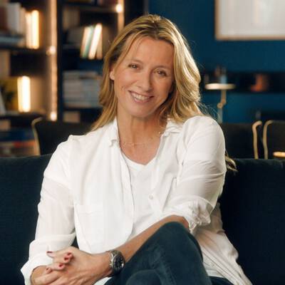 A woman with long, blonde hair smiles while sitting comfortably on a couch, wearing a white shirt and jeans. The background features a modern, stylish interior.