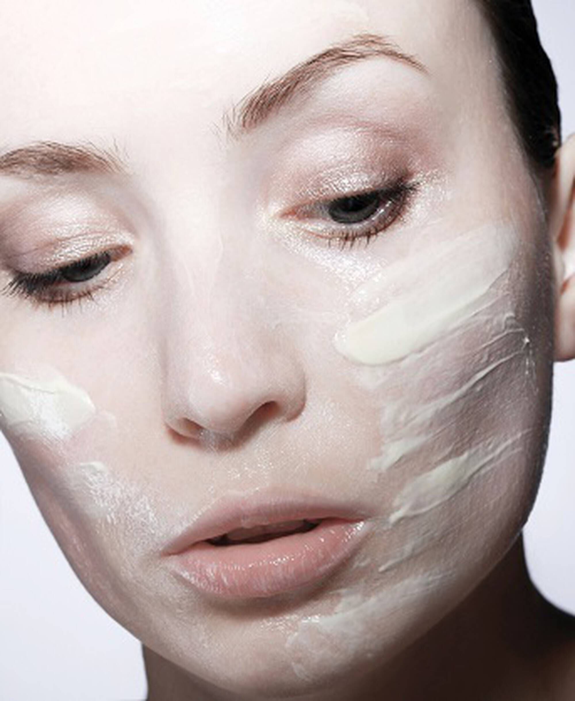 Femme avec un masque facial applique, regard pensif, peau lisse et lumineuse, fond clair.