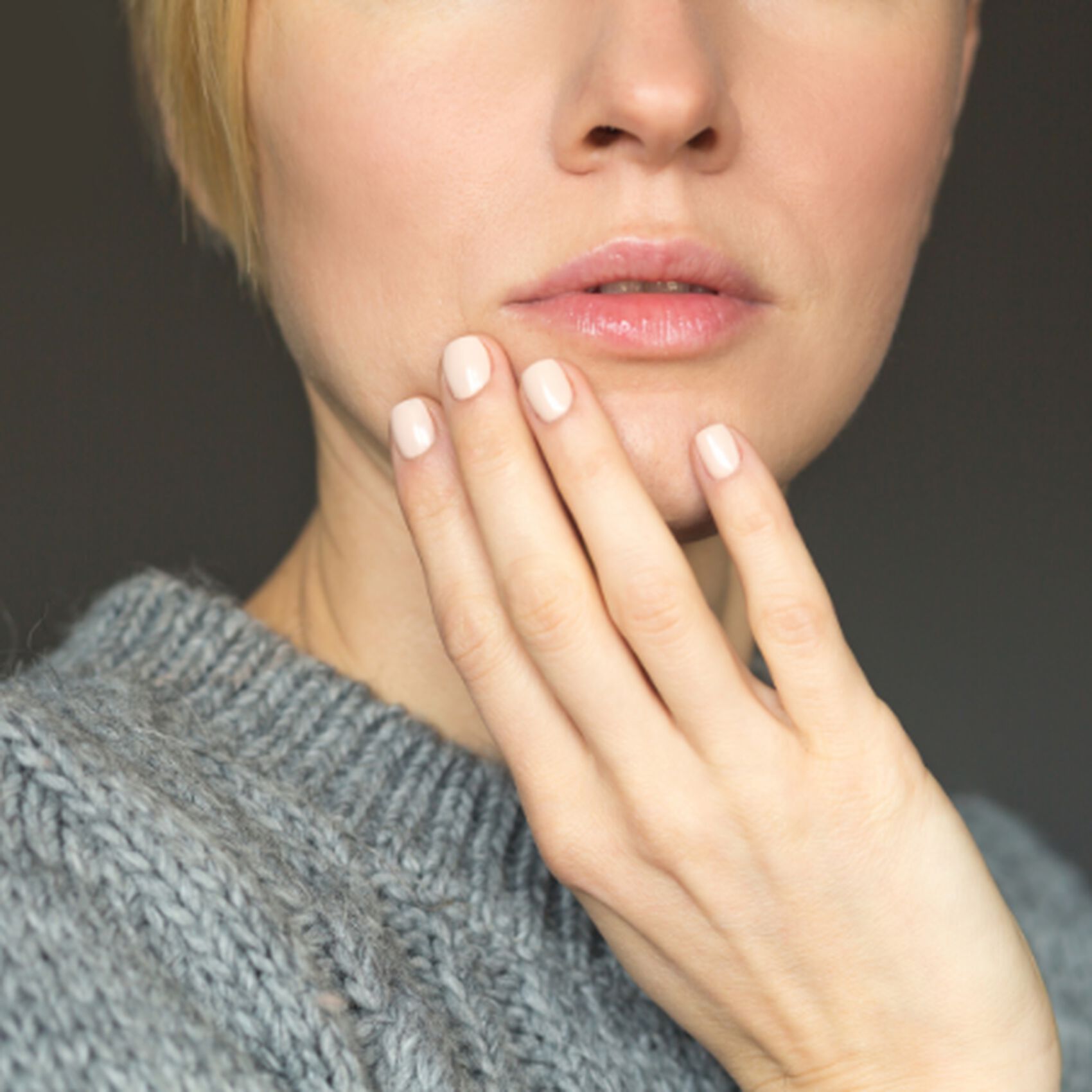 Femme aux cheveux blonds, main posee sur le visage, portant un pull en laine grise, regard pensif sur fond neutre.