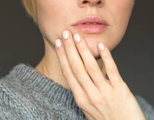 Femme aux cheveux blonds, main posee sur le visage, portant un pull en laine grise, regard pensif sur fond neutre.