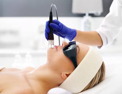 Une femme en train de recevoir un traitement facial avec un appareil, portant des lunettes de protection, pendant qu'un professionnel utilise un dispositif de soin.