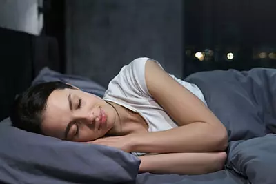 Une femme dort paisiblement dans un lit, souriante, avec un eclairage doux de la nuit a travers la fenetre.