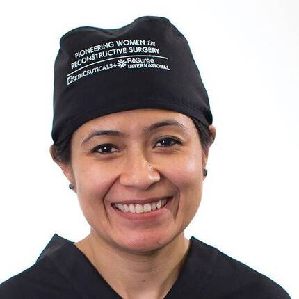 Une femme souriante portant un chapeau noir avec un texte, vetue d'une blouse, se tient devant un fond blanc.