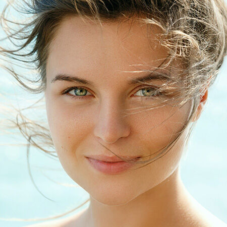 Femme au visage souriant, les cheveux au vent, avec des yeux verts et un arriere-plan flou de mer.