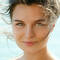 Femme au visage souriant, les cheveux au vent, avec des yeux verts et un arriere-plan flou de mer.
