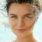 Femme au visage souriant, les cheveux au vent, avec des yeux verts et un arriere-plan flou de mer.