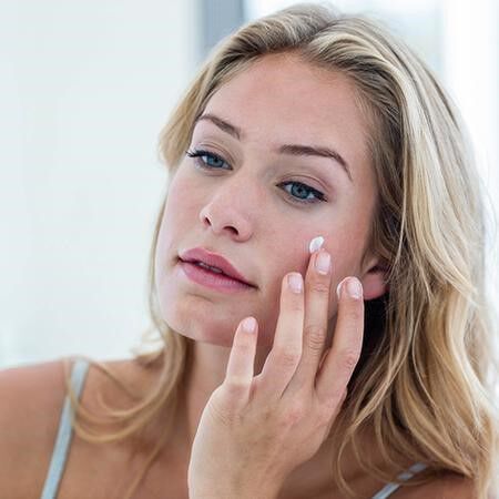 Une femme appliquant une creme sur son visage devant un miroir.