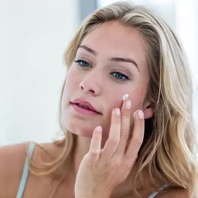 Une femme appliquant une creme sur son visage devant un miroir.