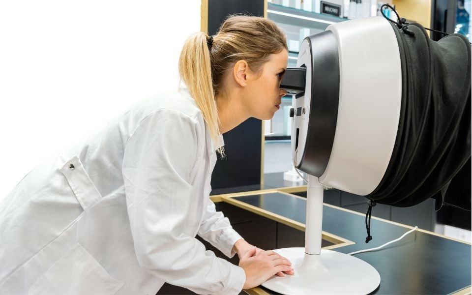 Une femme en blouse blanche se penche vers un appareil d'examen oculaire dans un environnement medical.