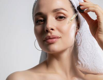 Femme appliquant un serum sur son visage, portant une serviette sur les cheveux et des boucles d'oreilles.