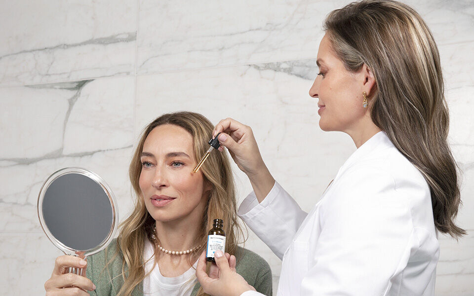 Une femme se regarde dans un miroir pendant qu'une autre personne applique un serum sur son visage.