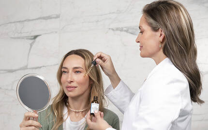 Une femme se regarde dans un miroir pendant qu'une autre personne applique un serum sur son visage.