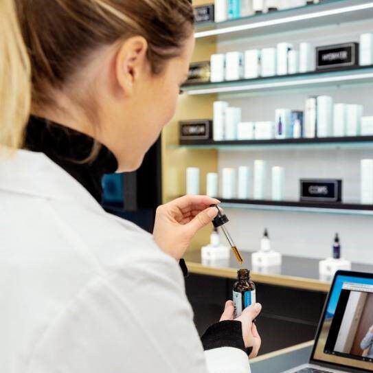 Une femme en blouse blanche utilise un compte-gouttes pour prelever un serum, avec des produits de soin de la peau en arriere-plan.