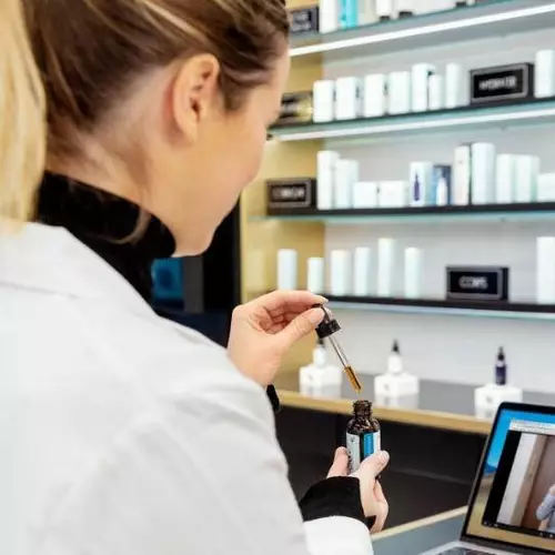 Une femme en blouse blanche utilise un compte-gouttes pour prelever un serum, avec des produits de soin de la peau en arriere-plan.