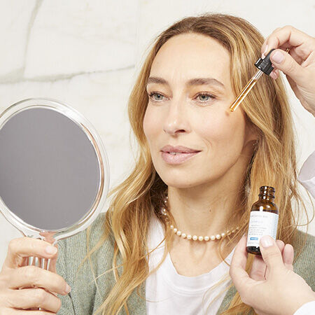 Femme souriante tenant un miroir et appliquant un serum sur son visage, avec une main qui utilise un compte-gouttes.