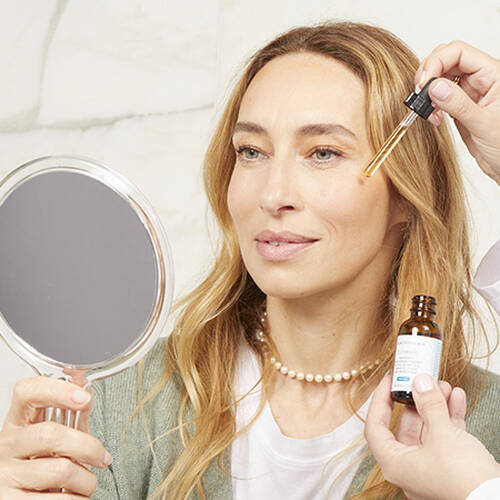 Femme souriante tenant un miroir et appliquant un serum sur son visage, avec une main qui utilise un compte-gouttes.