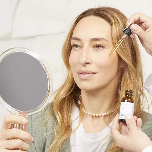 Femme souriante tenant un miroir et appliquant un serum sur son visage, avec une main qui utilise un compte-gouttes.