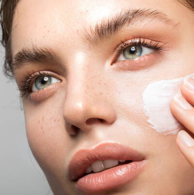 Femme en train d'appliquer une creme sur son visage, mettant en valeur sa peau eclatante et ses yeux clairs.