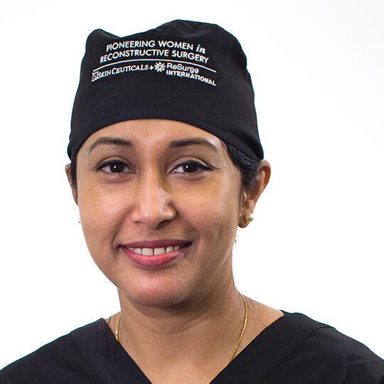 Une femme portant un uniforme medical et un bonnet, souriante, se tenant devant un fond blanc.