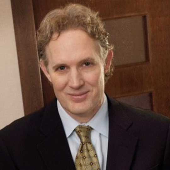 A smiling man with curly hair in a dark suit and tie, standing against a wooden background, presenting a professional demeanor.